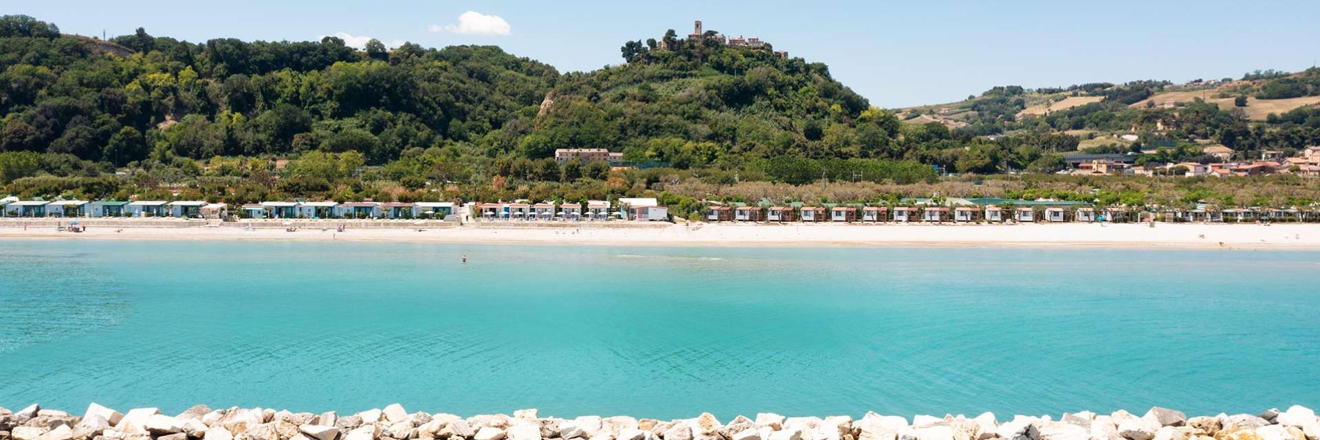 Beach with colorful cabins, crystal-clear sea, and green hills in the background.