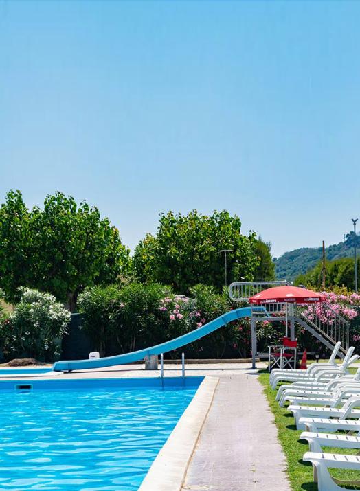 Piscina all'aperto con scivolo e lettini bianchi, circondata da alberi e fiori.