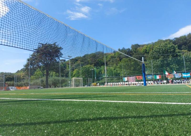 Campo sportivo all'aperto con rete e bandiere, circondato da alberi.